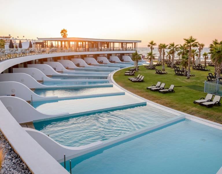Lyttos Gallery resort pool area at sunset with infinity pools, sun loungers, palm trees, and ocean view.