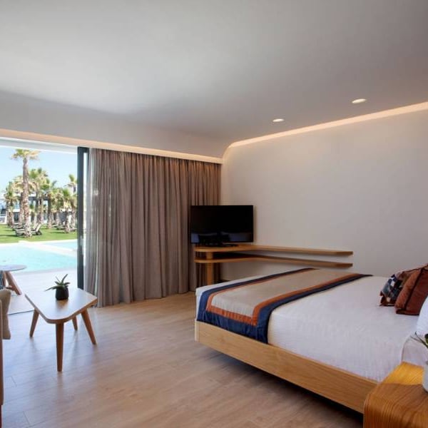 Hotel room at Lyttos Gallery with a bed, sofa, and TV, overlooking palm trees through a glass door.