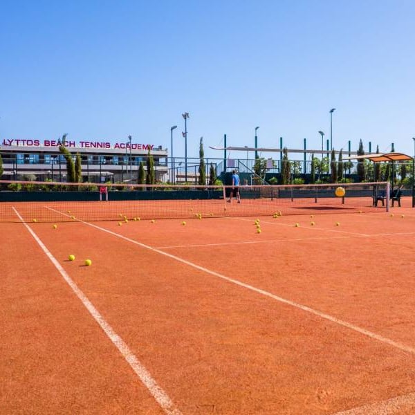 Clay tennis court at Lyttos Beach Tennis Academy with tennis balls scattered on the ground and a net in the centre.
