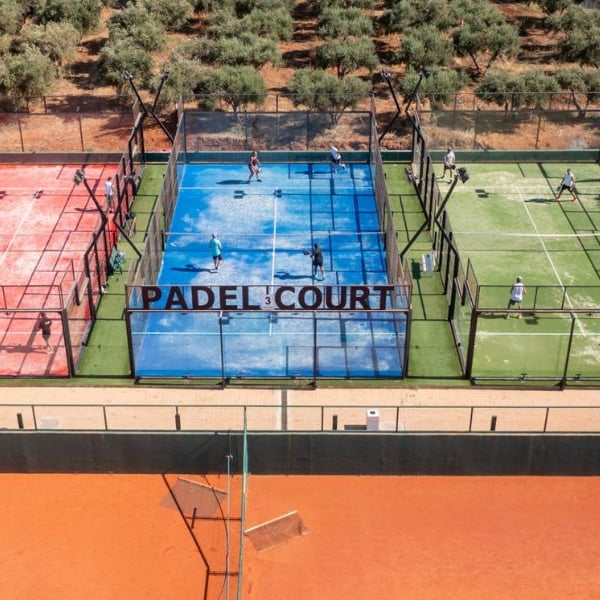 Aerial view of Lyttos Mare's padel courts with players, featuring red, blue, and green courts surrounded by an olive grove.