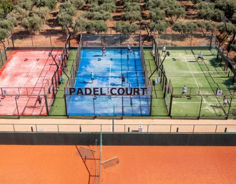 Aerial view of Lyttos Mare's padel courts with players, featuring red, blue, and green courts surrounded by an olive grove.