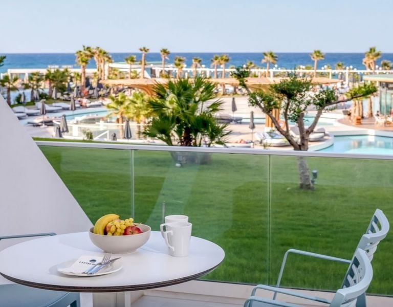 View from a balcony at Lyttos Mare, showing the pool area, palm trees, green lawns, and the sea in the background.
