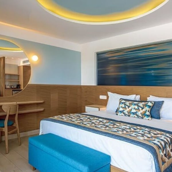 Hotel room at Lyttos Beach with a large bed, blue and beige linens, a wood desk with a round mirror, and ambient lighting.