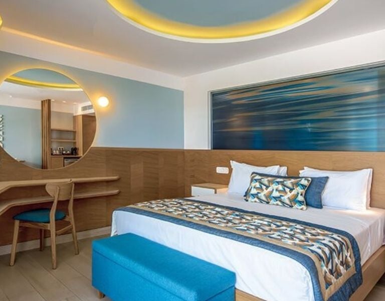 Lyttos Mare hotel bedroom with double bed, geometric bedspread, round mirror, and wooden furnishings.