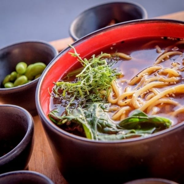 Asian noodle soup in a black and red bowl with herbs, surrounded by bowls of edamame and condiments on a wooden tray.