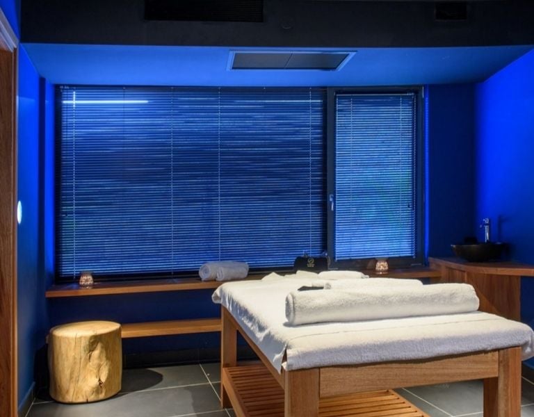 Lyttos Mare spa room with massage table, wooden bench, candles, and deep blue walls for a calming atmosphere.