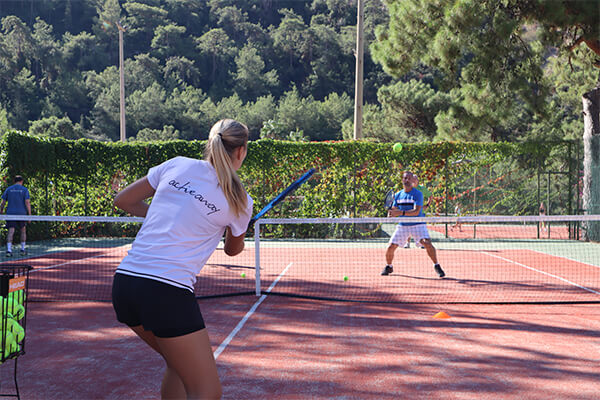 Marie-Lou playing tennis on a clay court, hitting the ball to an opponent in front of lush greenery and tall trees.