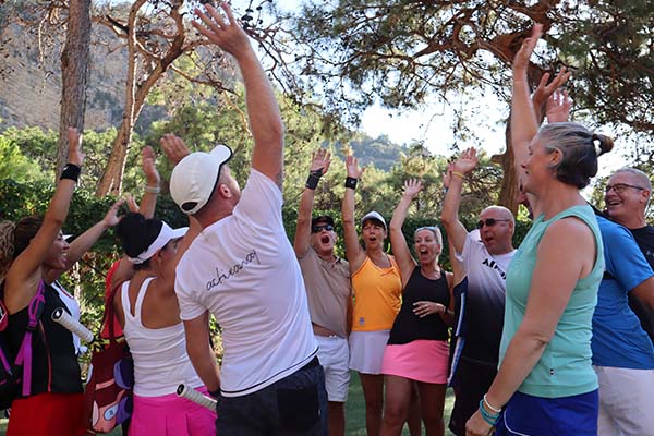Group of people in outdoor fitness gear raising their arms together in a joyful team-building activity under trees.