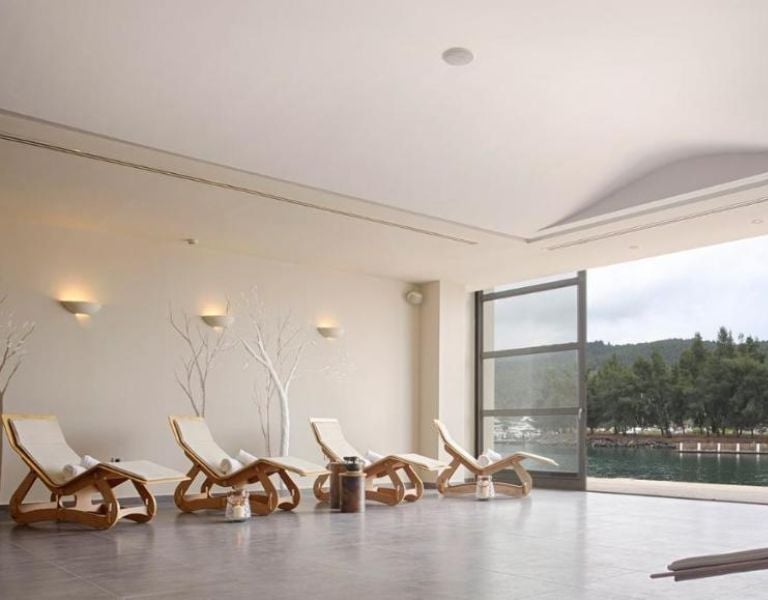 Indoor lounge of Meliton Gallery with wooden reclining chairs and a large window showing a lake and greenery.