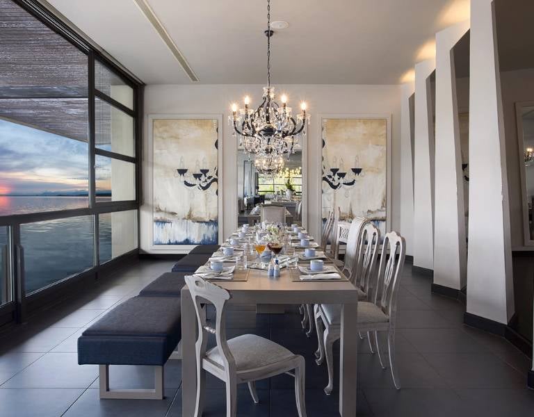 Dining room at Meliton Gallery Restaurant, with a long table set, chandeliers, large windows showing a sunset view, and modern art on the walls.