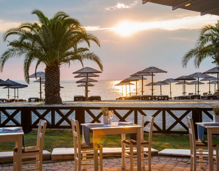 Seafront dining area at Meliton Gallery with wooden tables, chairs, straw parasols, and a sunset over the ocean.