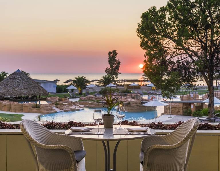 Dinner table set for two overlooking a pool at Meliton Gallery, with a sunset view over the sea and surrounding palm trees.