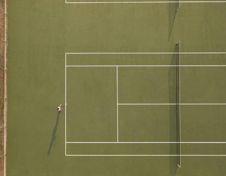 Aerial view of a tennis court at Meliton Gallery with a single tennis player preparing to serve.