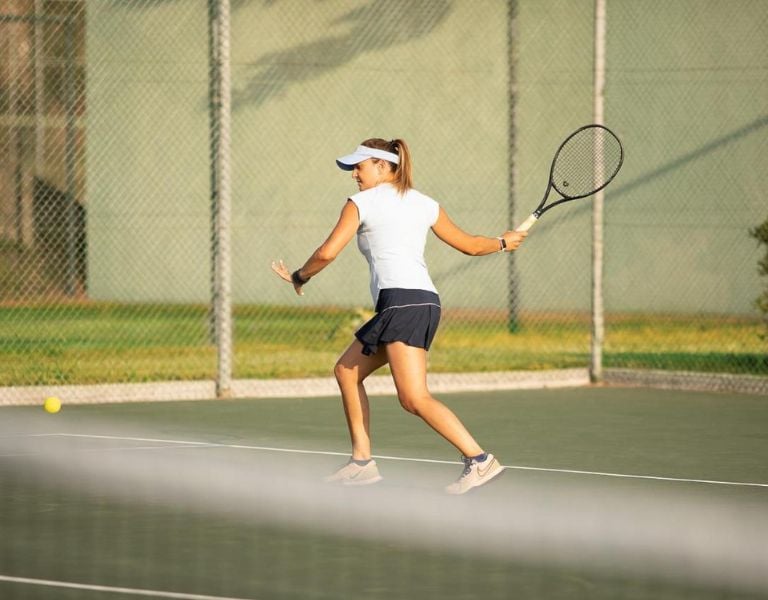 Female tennis player prepares for a forehand shot on an outdoor court, wearing a visor and sports attire, with sunlight highlighting the scene.