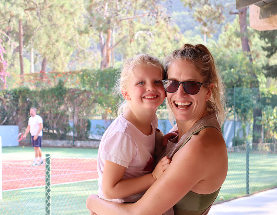 Mother and daughter smiling together at a tennis court with a man playing in the background, showcasing a family-friendly atmosphere.