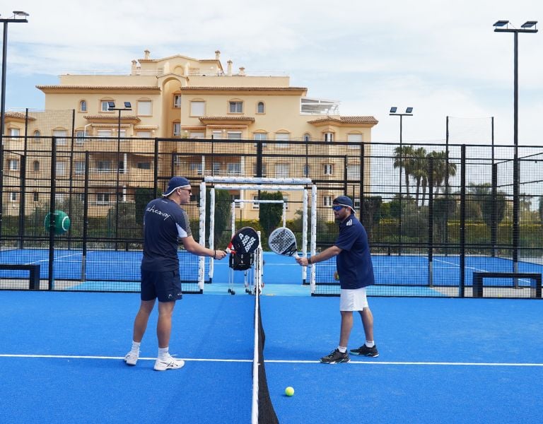 Two coaches on a blue padel court at Oliva Nova Beach Resort, holding racquets and having a discussion with luxury apartments in the background.