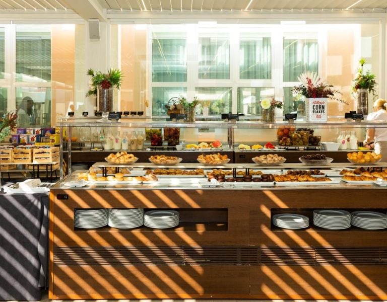 Buffet at Oliva Nova Padel with pastries, fruits, and breakfast items in a bright dining area with glass windows and potted plants.