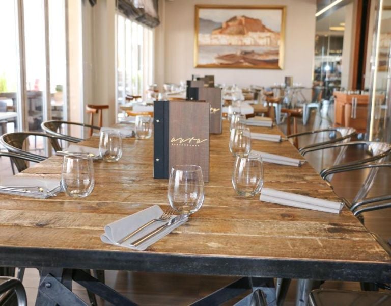 Oliva Nova dining area with a long wooden table, wine glasses, napkins, and contemporary chairs, framed by a large painting.