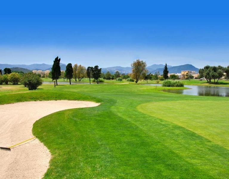 Golf course at Oliva Nova, featuring a sand bunker, fairways, a water hazard, and mountains in the backdrop.