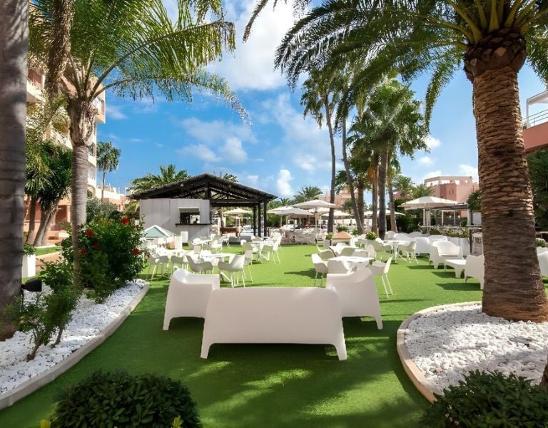 Outdoor lounge at Oliva Nova with white furniture, palm trees, and green artificial turf.