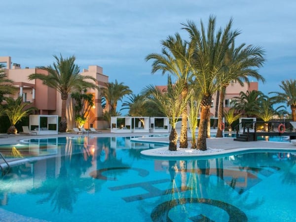 Outdoor pool at Oliva Nova Resort with palm trees, modern cabanas, and dusk lighting reflecting on the water surface.