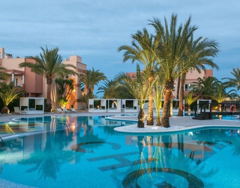 Outdoor pool at Oliva Nova Resort with palm trees, modern cabanas, and dusk lighting reflecting on the water surface.