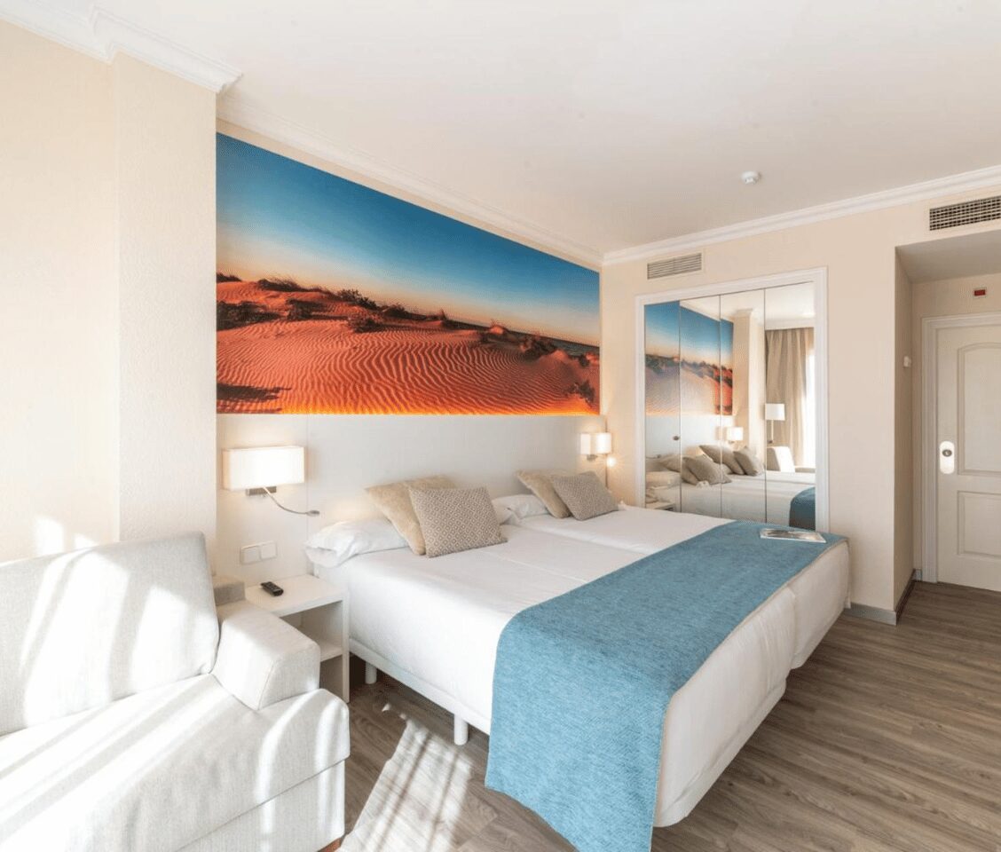 Oliva Nova Golf Resort room with two double beds, beach mural, grey sofa, and modern decor.