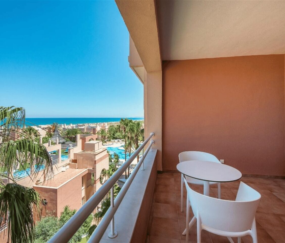 Balcony of an Oliva Nova sea view room with white table and chairs, overlooking the resort, palm trees, and ocean.