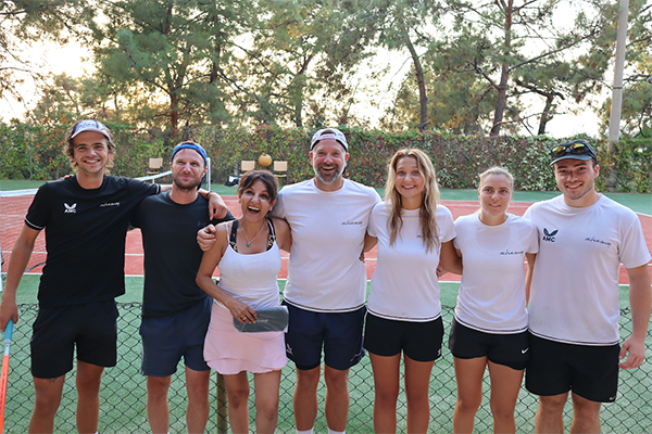 Seven smiling team members posing on a tennis court with trees in the background, wearing sports attire.