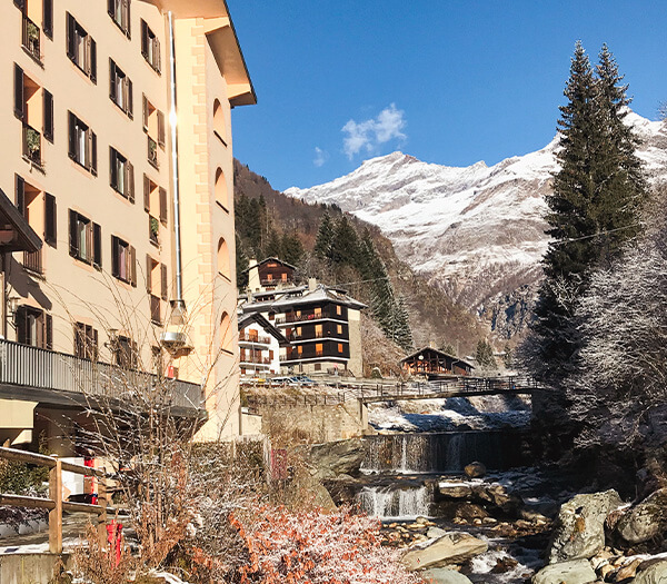 Cristallo Hotel in Italy with traditional wooden buildings, a flowing stream, snow-dusted trees, and the snow-capped Alps under a blue sky.