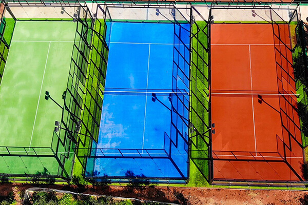 Aerial view of three distinctive padel tennis courts in green, blue, and red, all enclosed by wire fences and surrounded by green landscape.