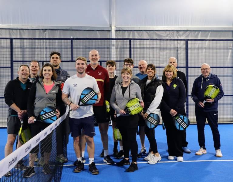 Group of men and women holding padel rackets on a blue indoor padel court, posing in front of the net.