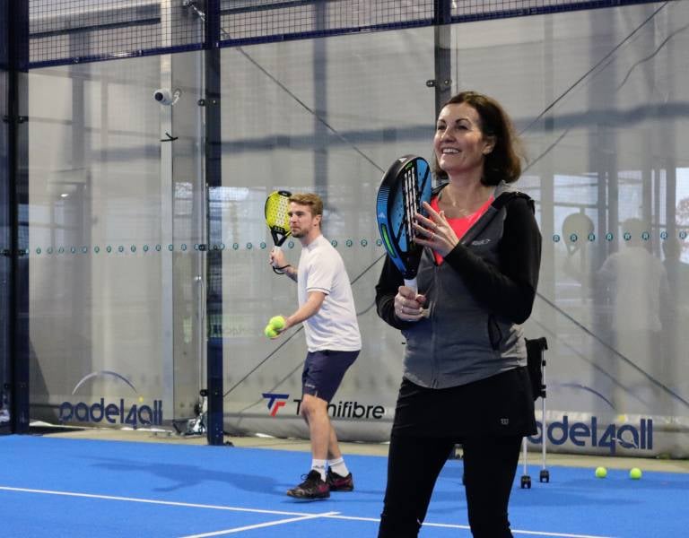 A smiling woman with a padel racket in an indoor court, coaching session under the padel4all brand, man in the background preparing to play.