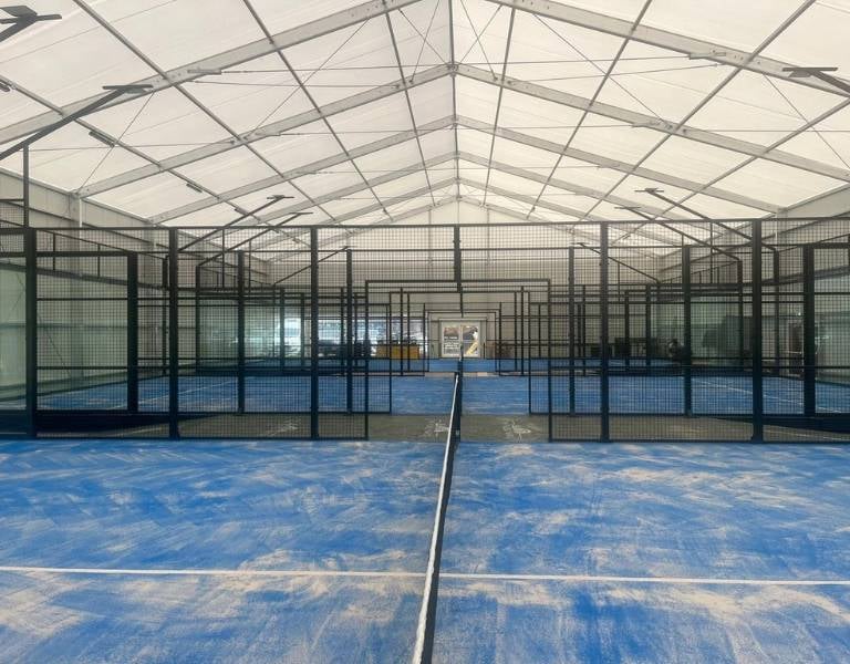 Indoor padel facility with multiple blue courts and black mesh partitions under a white canopy.