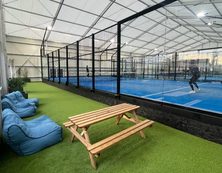 Indoor padel courts with surrounding glass walls, players in action, bean bag seating, and a picnic table on artificial grass.