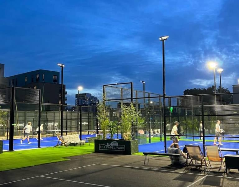 Evening view of The Padel Yard's outdoor courts with players and an urban backdrop, illuminated by lights.