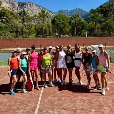 Group of female tennis players on a clay court, wearing colourful outfits and holding rackets, with mountains in the background.