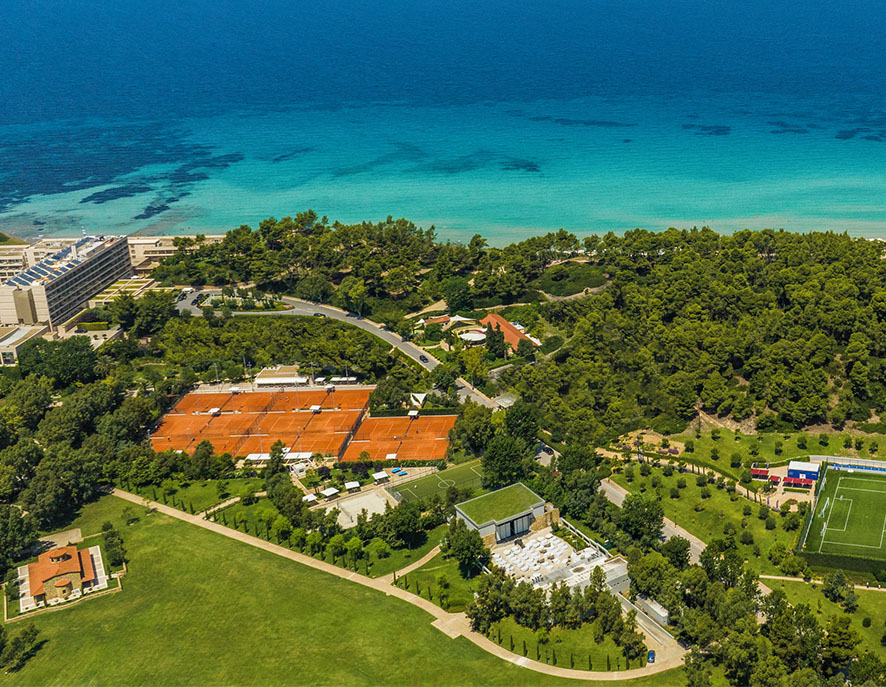 Aerial view of Sani Resort in Greece, showcasing lush greenery, turquoise sea, tennis courts, and a football field.