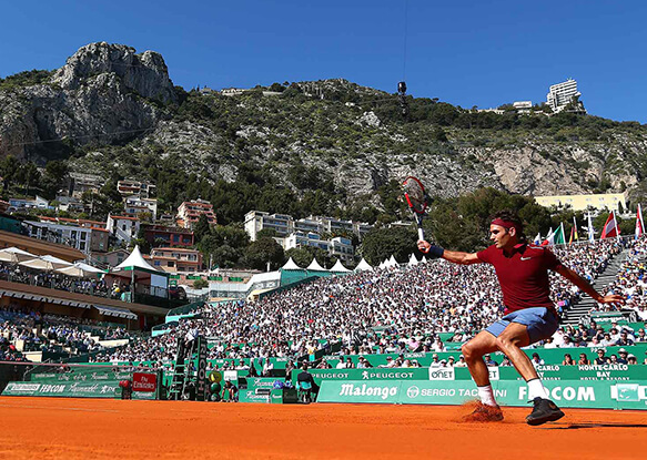Tennis player on clay court at Monte-Carlo Country Club, with a crowd in the stands and cliffs in the background.