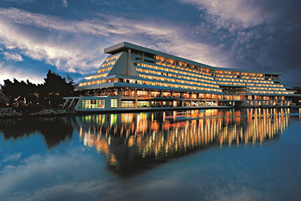 Night-time view of the illuminated Porto Carras Grand Resort, with its reflection in the water, against a dark evening sky.