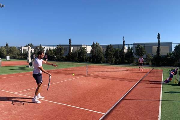Two tennis players on a clay court warming up, with one about to hit the ball while the other waits on the opposite side. Surrounded by green trees and a clear blue sky.