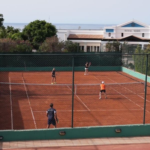 Players on a clay tennis court at Royal Mare hotel with lush greenery and hotel building in the background.
