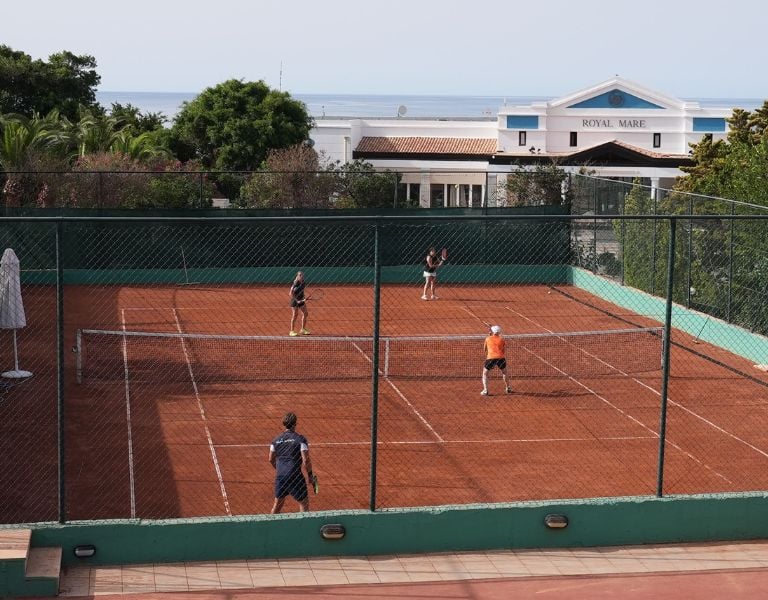 Players on a clay tennis court at Royal Mare hotel with lush greenery and hotel building in the background.