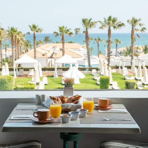 Breakfast table with croissants, orange juice, and coffee, overlooking a garden with palm trees and sea view at Royal Mare.