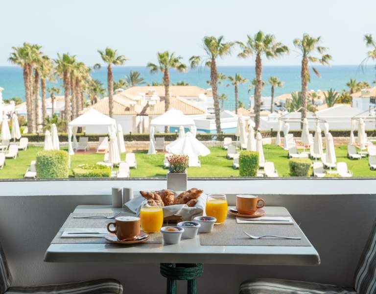 Breakfast table with croissants, orange juice, and coffee, overlooking a garden with palm trees and sea view at Royal Mare.