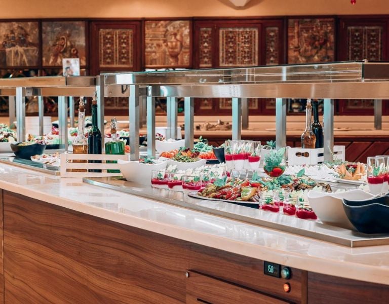 Luxurious buffet at Royal Mare Gallery with gourmet dishes, salads, drinks, and elegant presentation on a wooden counter with stainless steel warming lamps.