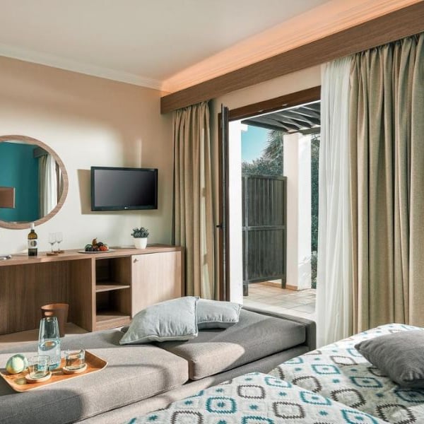 Modern hotel room at Royal Mare with a bed, patterned bedding, wooden console, round mirror, flat-screen TV, and balcony door open to natural light.