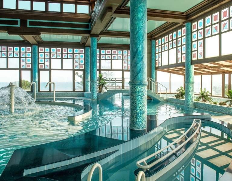 Luxurious indoor spa area at Royal Mare Gallery, featuring a large pool with mosaic columns and panoramic windows with views outside.
