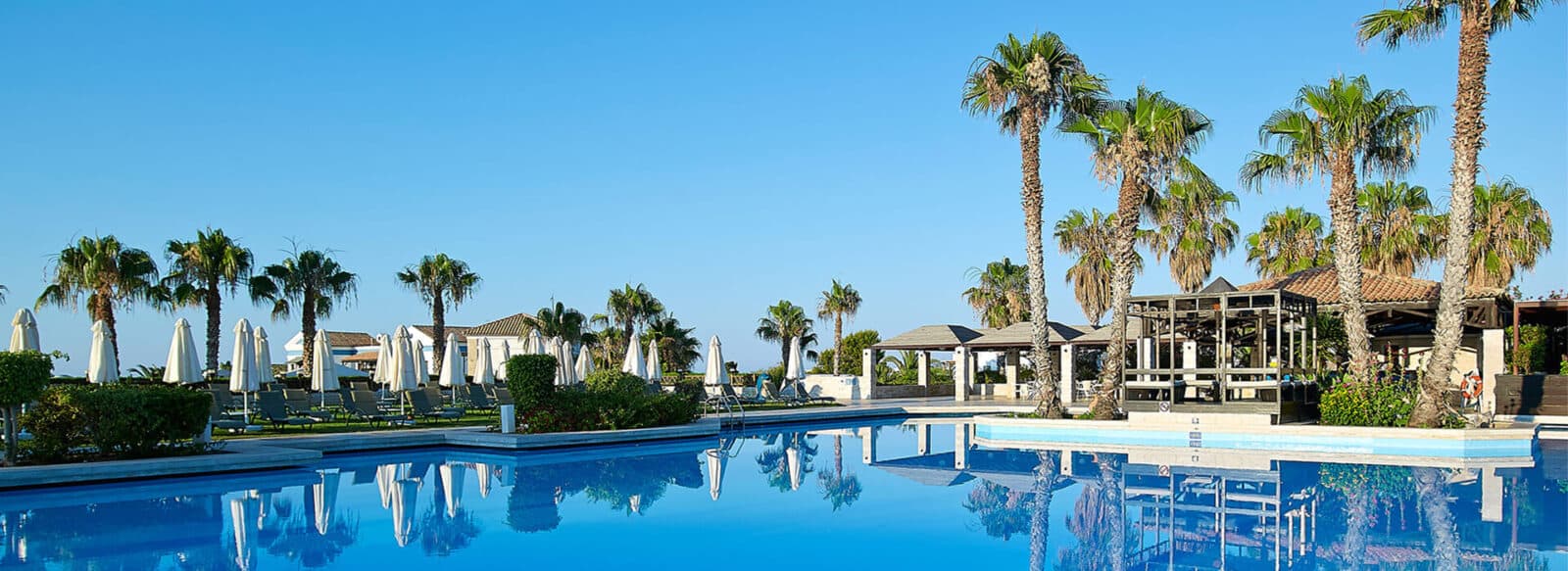 Royal Mare resort pool with surrounding palm trees and sun loungers under a clear blue sky.