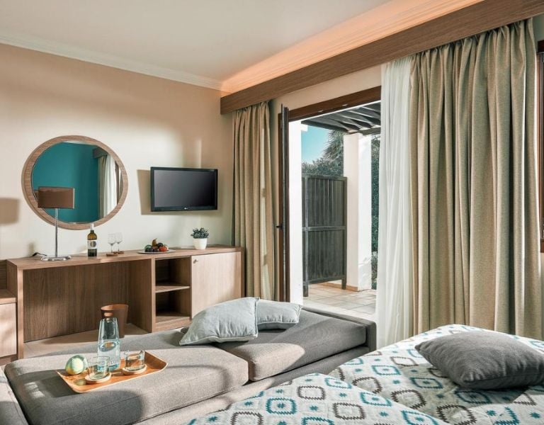 Luxurious hotel room at Royal Mare with a grey chaise lounge, flat-screen TV, round mirror, and access to private terrace through sliding doors.